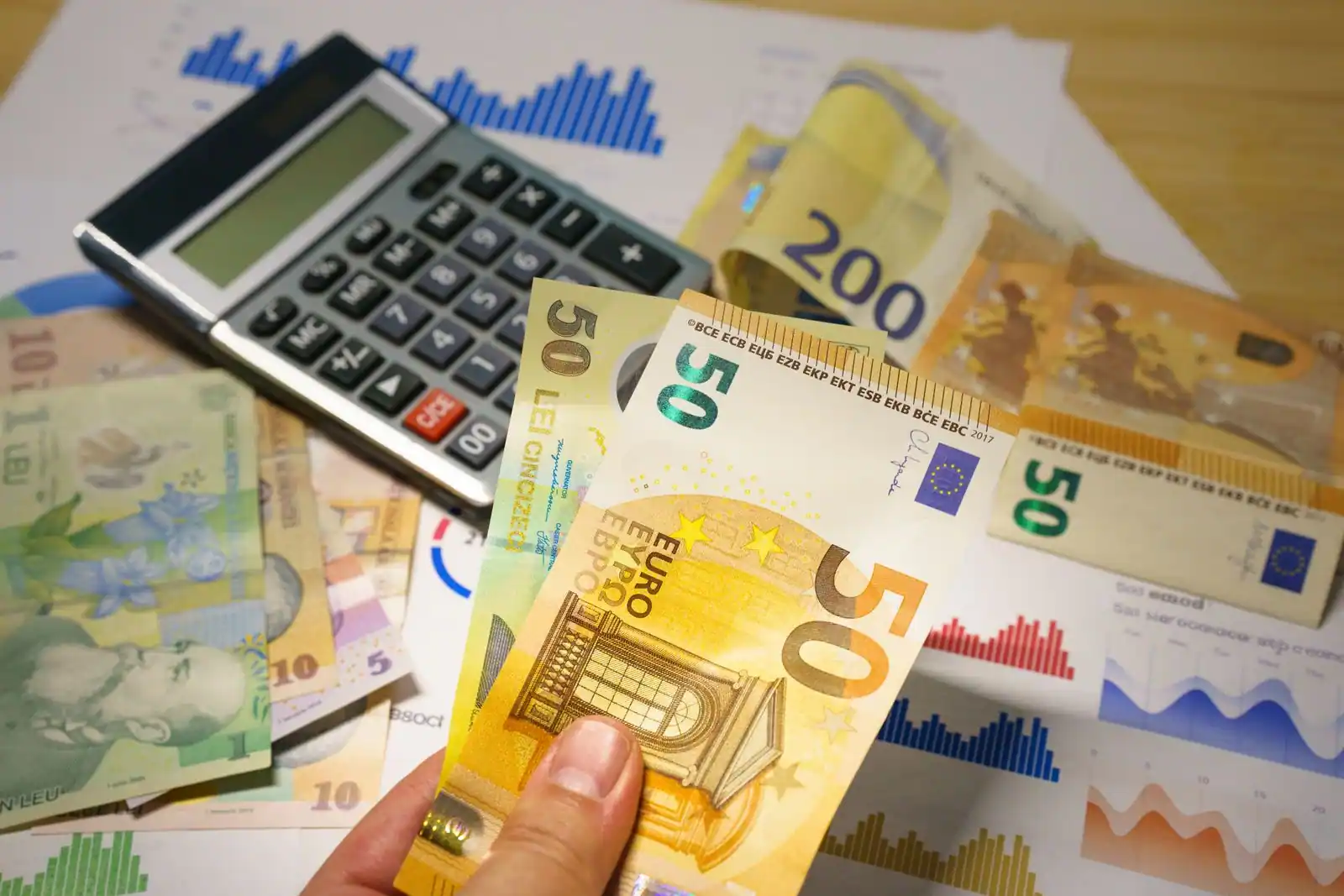 Calculatrice et billets de banque, dont des coupures en euros, posés sur des graphiques financiers imprimés.