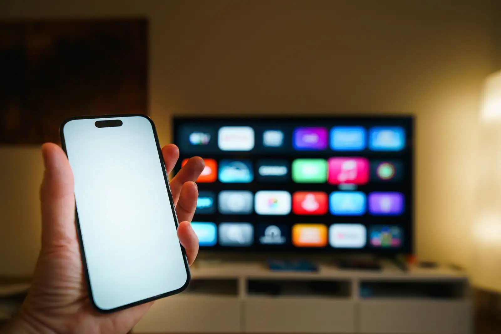 Main tenant un smartphone devant un téléviseur connecté affichant des applications, dans un salon.
