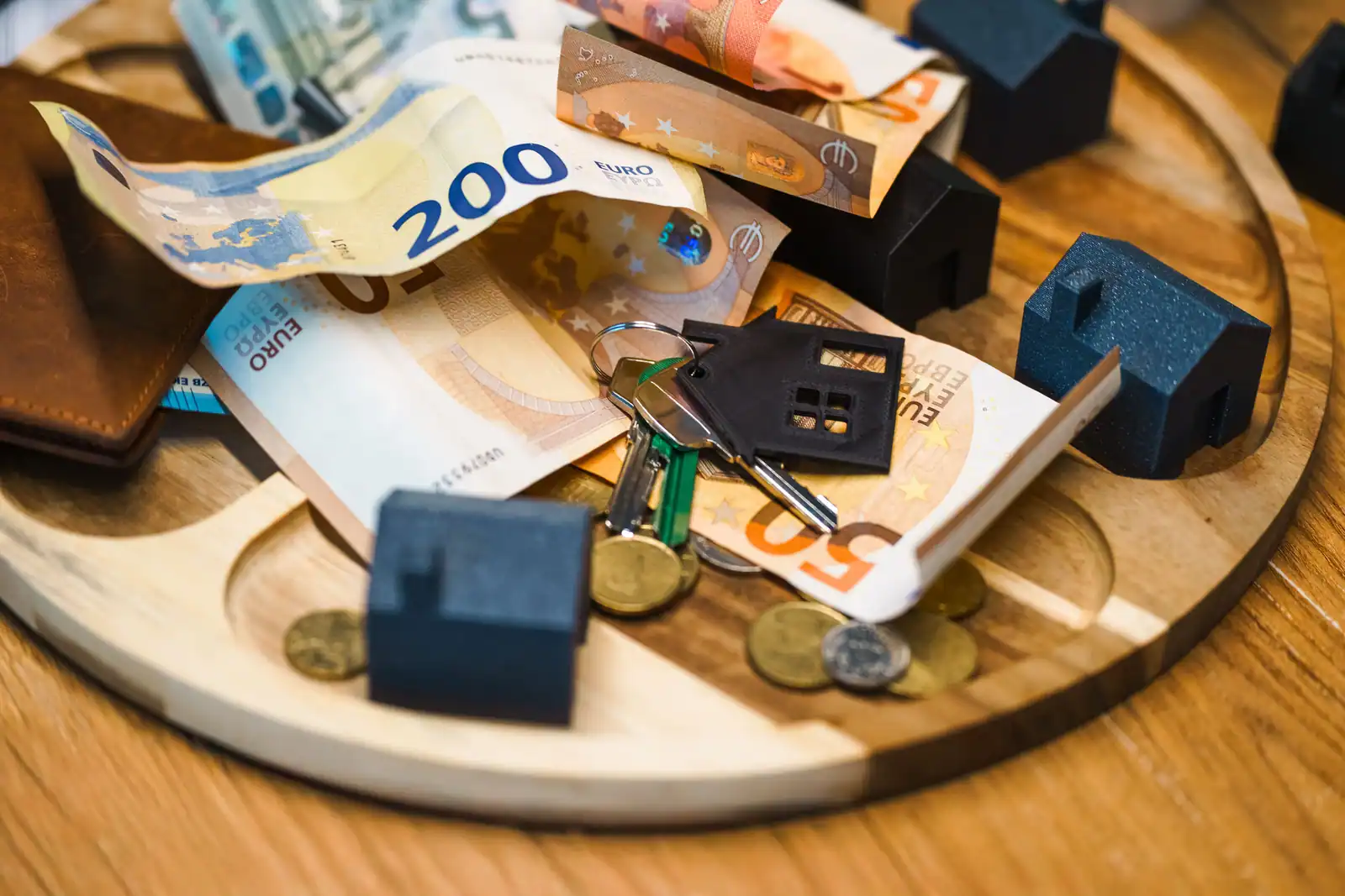 Billets en euros, clés de maison et petites maisons miniatures sur un plateau en bois, évoquant l'épargne en vue d'un projet immobilier