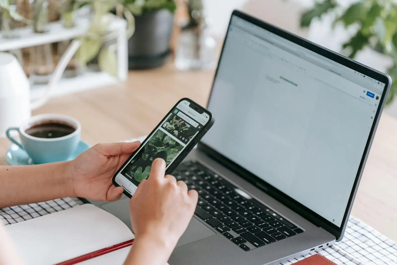 Personne tenant un smartphone devant un ordinateur portable ouvert, sur un bureau avec une tasse de café