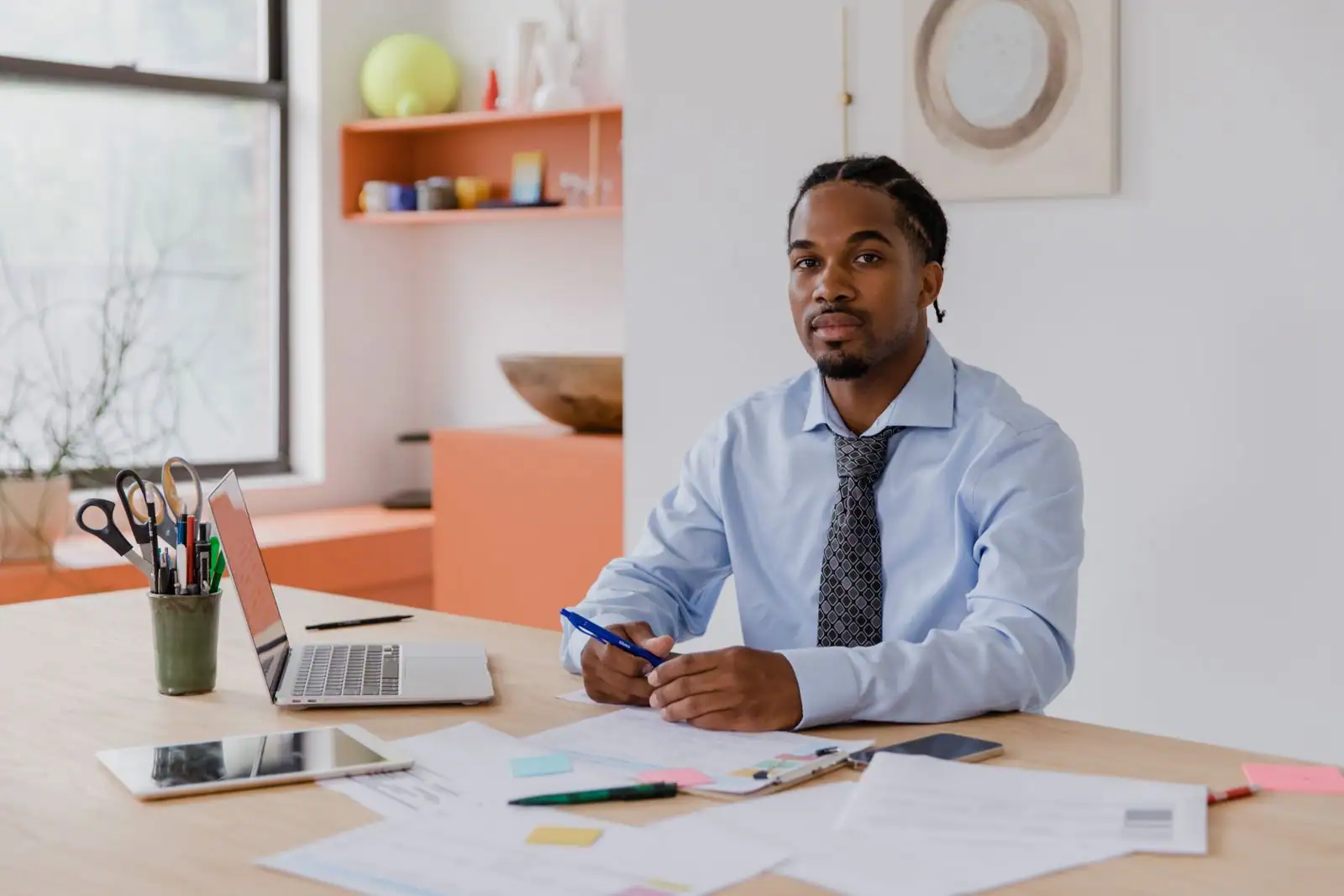 Jeune entrepreneur en chemise assis à son bureau, avec un ordinateur portable, une tablette et des documents de travail étalés