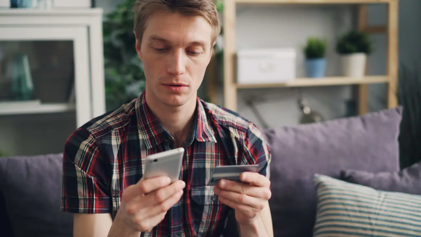 Homme assis dans un salon, son téléphone dans une main et sa carte bancaire dans l'autre.